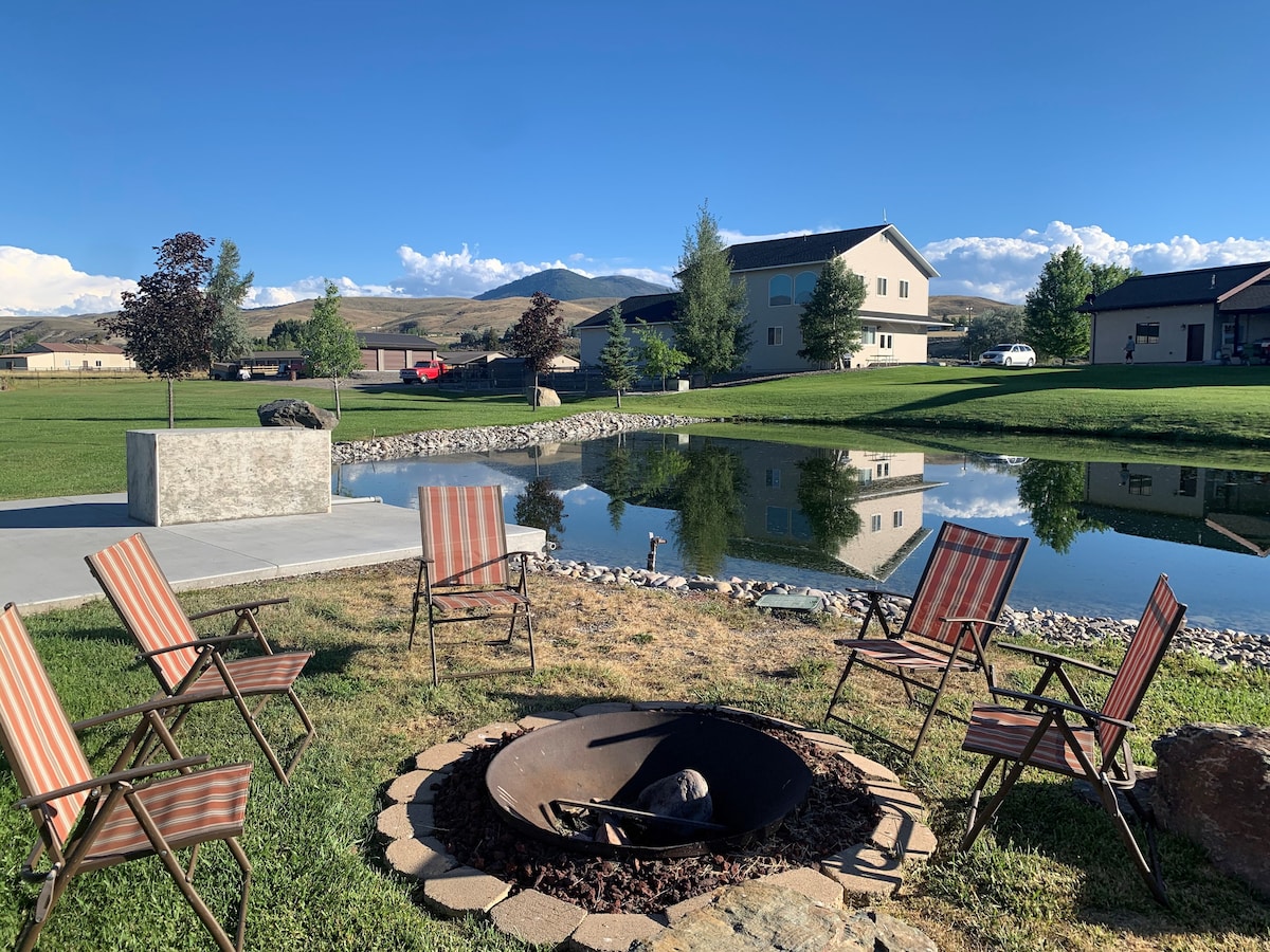 A serene outdoor space features a fire pit surrounded by six folding chairs on a grassy area. A pond reflects nearby trees and buildings, with mountain views in the background under a clear blue sky.