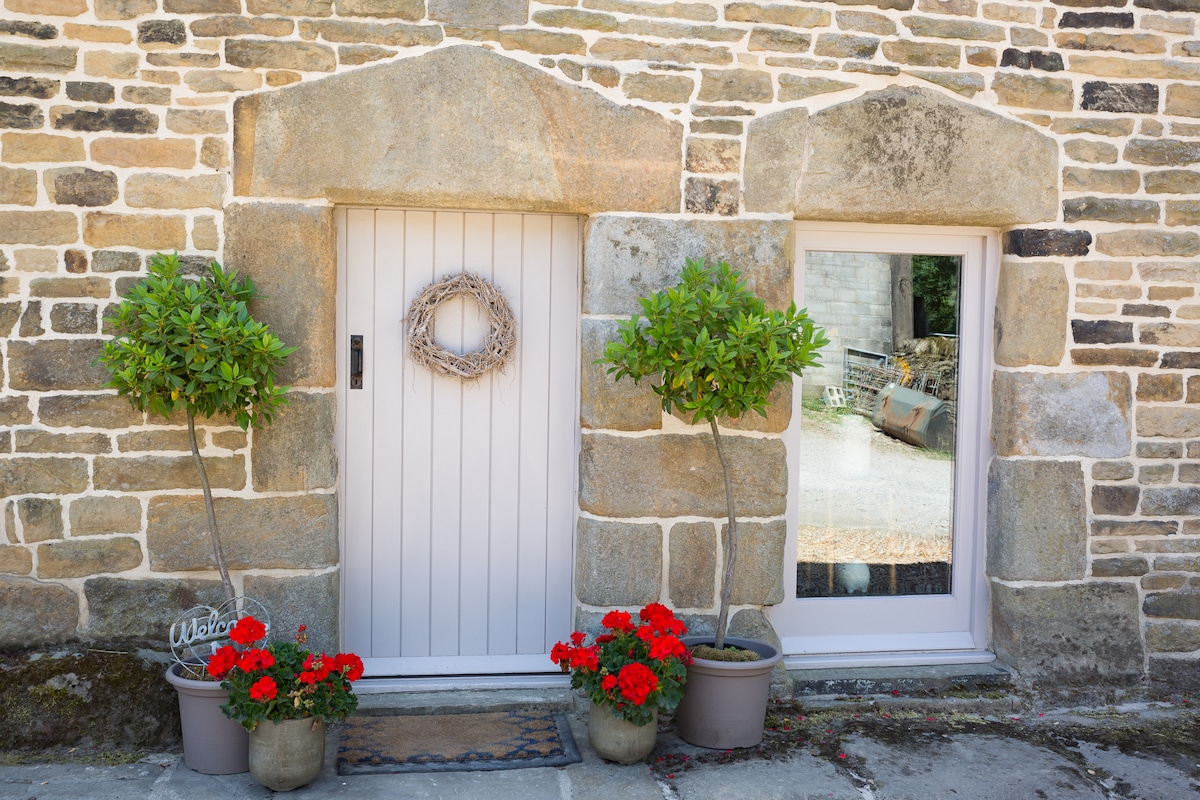An entrance is framed by textured stone walls, featuring a pale wooden door adorned with a circular wreath. Two potted plants flank the door, accompanied by vibrant red flowers in pots. A reflection of the outdoor space is visible in a nearby window.