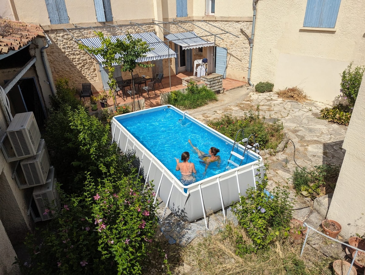An above-ground pool is seen in a private courtyard, surrounded by lush greenery and natural stone pathways. Two individuals are enjoying the water. A shaded seating area with striped awnings is visible in the background, adding to the inviting outdoor space.