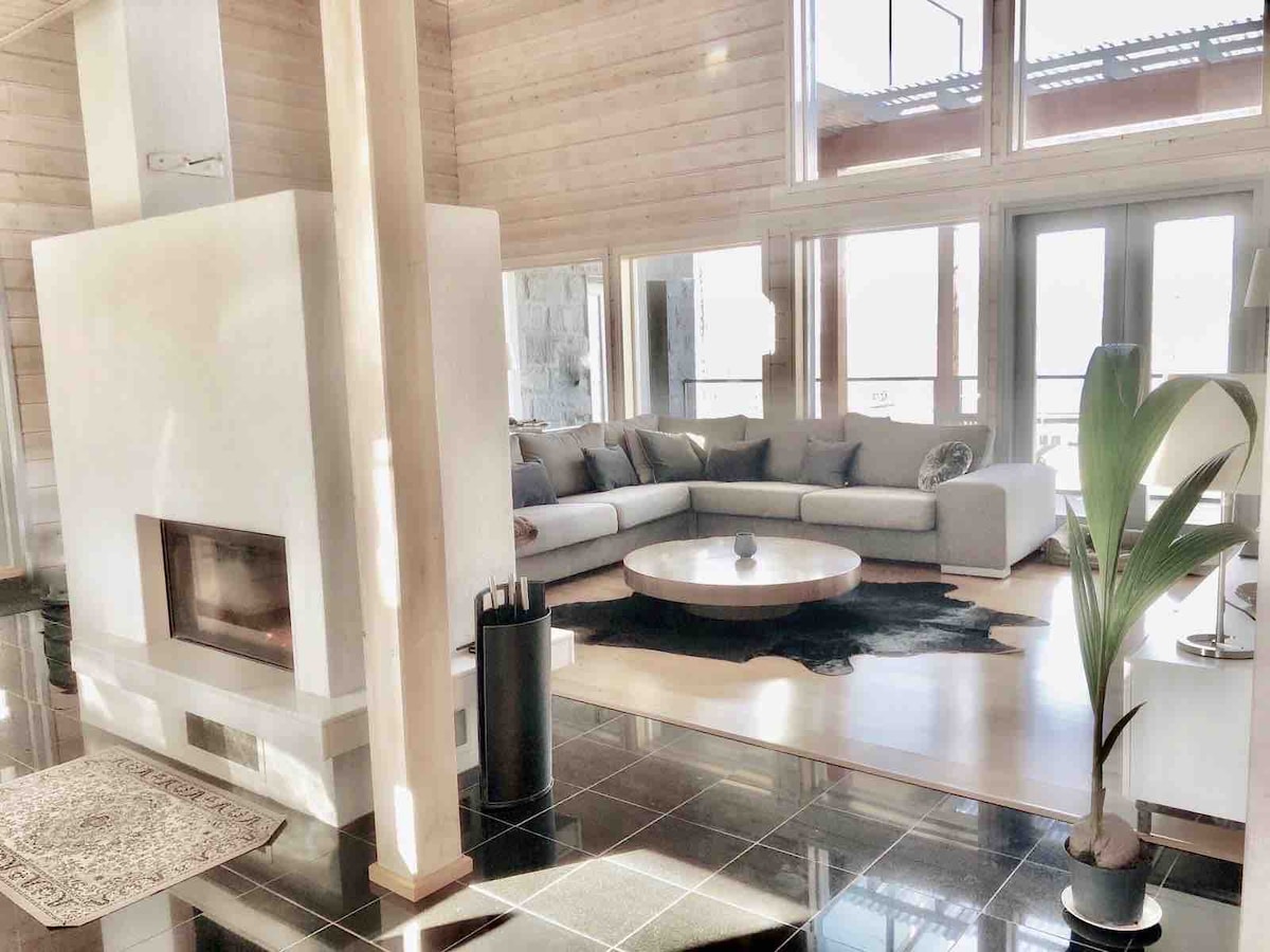 A spacious living area features a modern, curved sectional sofa arranged around a circular coffee table. A sleek fireplace is positioned against a textured wall, while large windows allow natural light to illuminate the room. A decorative rug enhances the floor space.