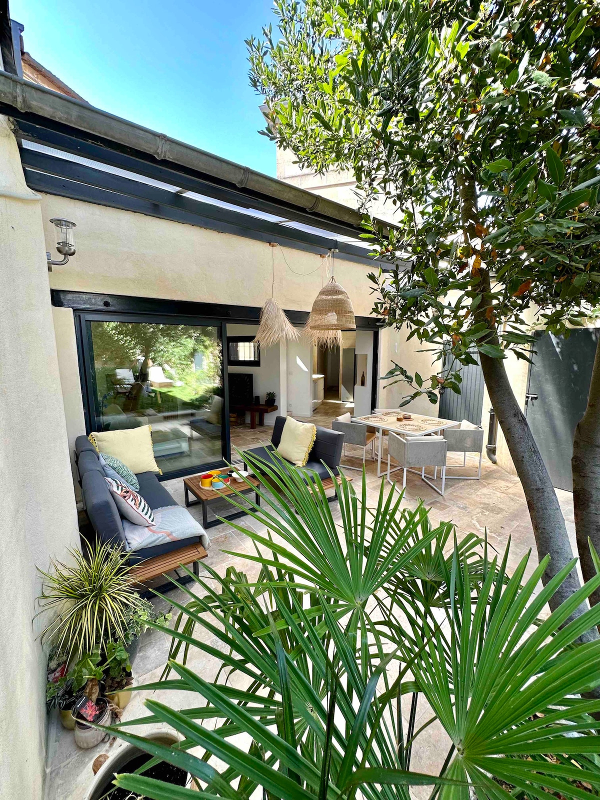 A peaceful outdoor area features comfortable seating arranged around a small table, surrounded by greenery. Large glass doors provide a view into the interior space, while a decorative hanging light adds charm. Sunlight filters through, enhancing the serene atmosphere.