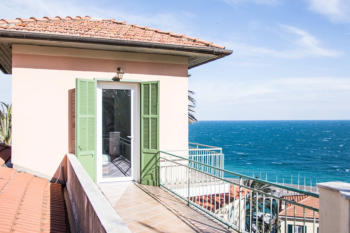 A private terrace with green shutters frames a sweeping view of the blue ocean. Sunlight reflects off the water, creating a serene atmosphere. The tiled floor extends towards the railing, emphasizing the outdoor space's openness and connection to the seaside.