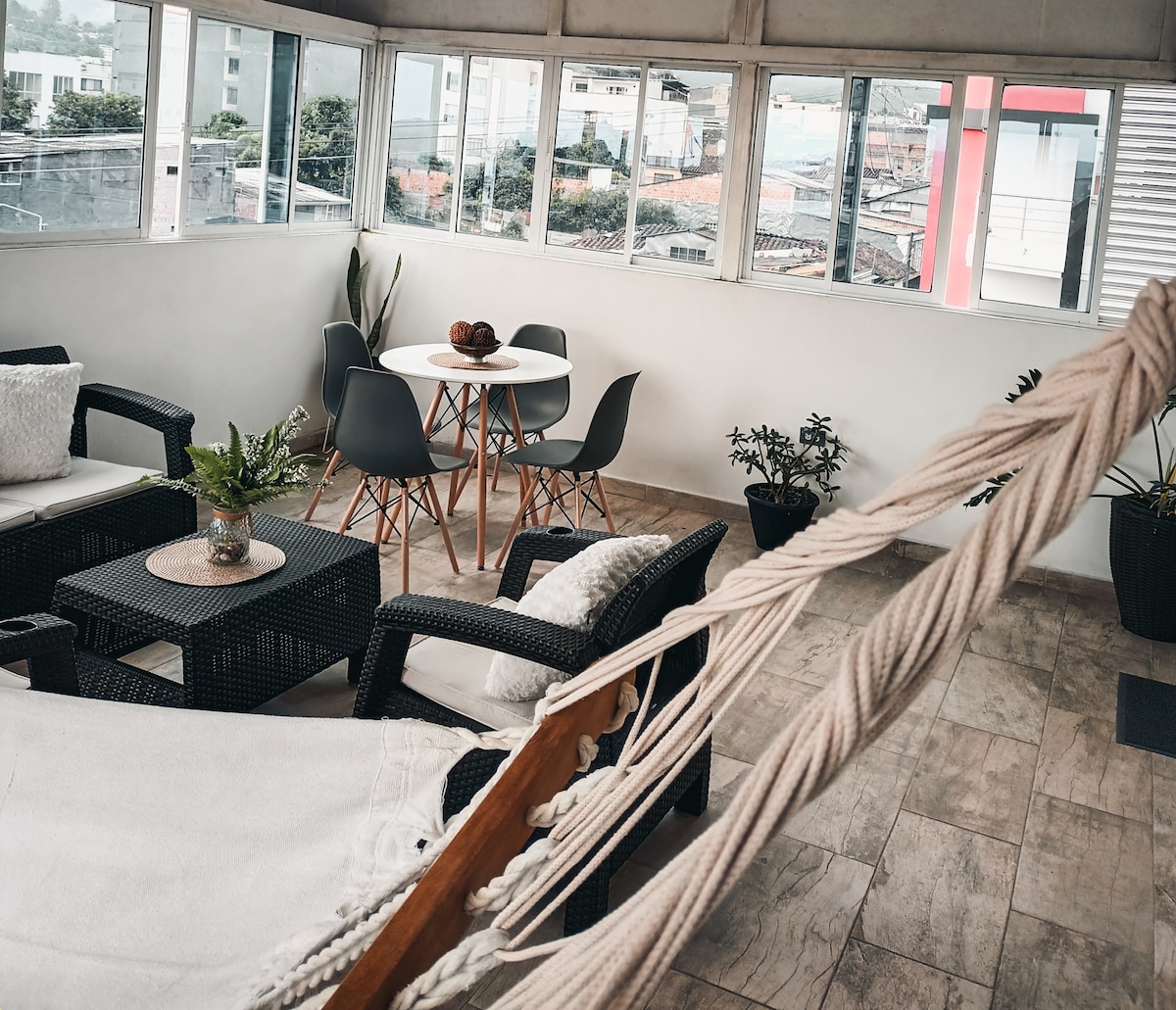 An inviting seating area features a hammock alongside wicker chairs and a small round dining table. Sunlight enters through large windows, illuminating the room and plants that add greenery to the space.