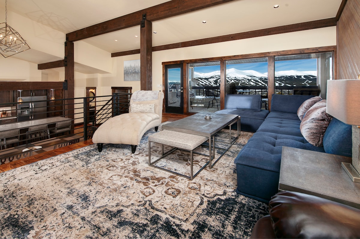 A spacious living area is highlighted by large windows offering views of the snow-capped mountains. A sectional sofa, plush chaise lounge, and an elegant coffee table are arranged on a patterned area rug. Warm wood accents complement the modern mountain decor.