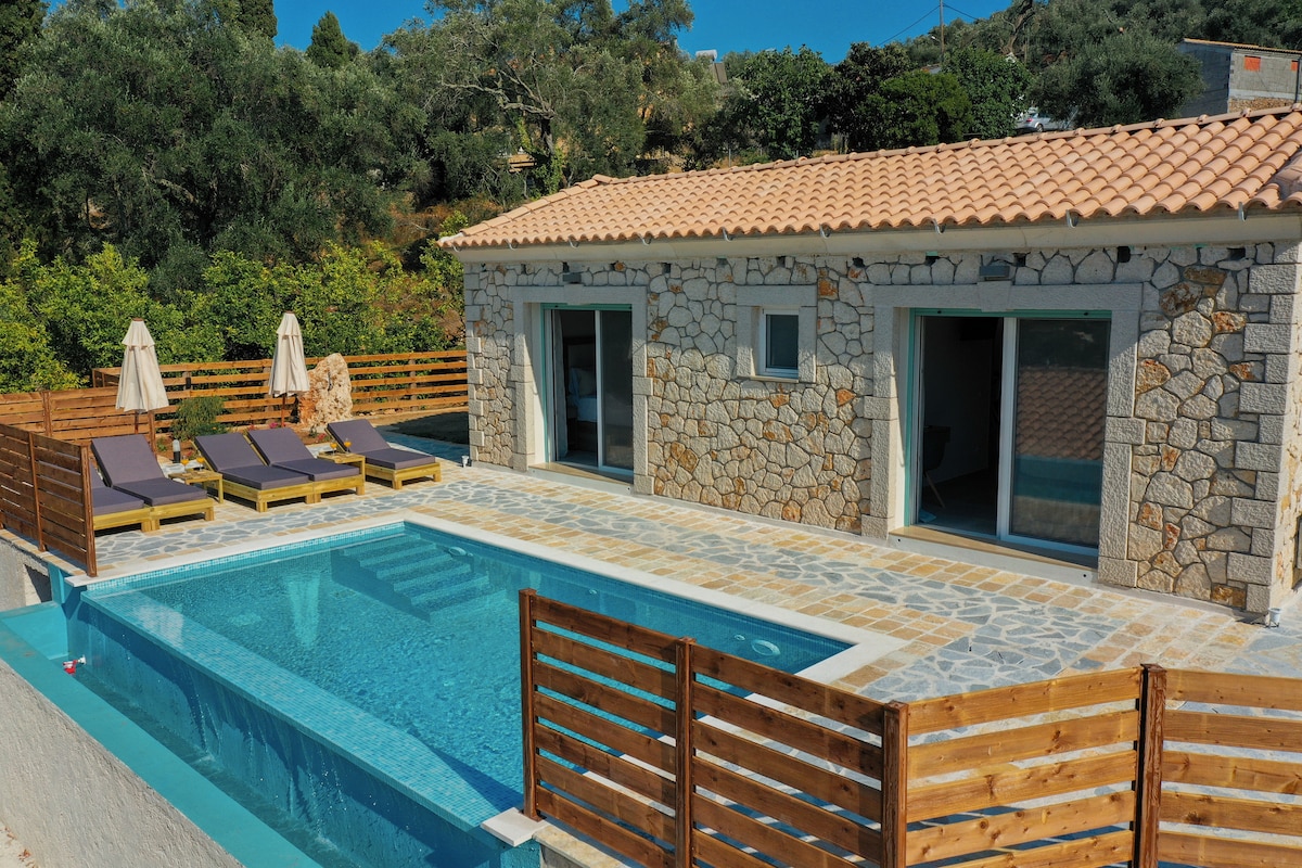 A sunlit private villa is displayed, featuring a serene pool surrounded by a stone patio. Comfortable lounge chairs are positioned alongside the pool, with two umbrellas providing shade. The villa's stone exterior complements the natural landscape, with oversized glass doors opening to the spacious interior.