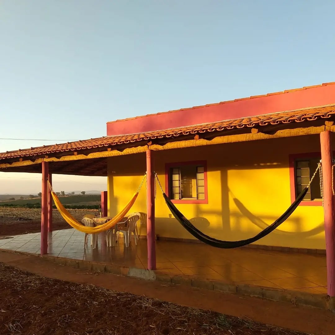 A vibrant yellow house is shown with a spacious covered porch. Two hammocks are suspended between pink columns, providing a relaxed seating option. White plastic chairs surround a small table, and large windows allow natural light to enter the living space.