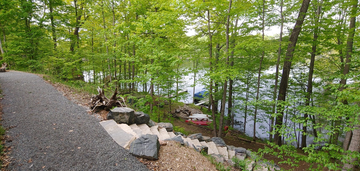 A stone path leads through lush greenery toward the water's edge. The clear lake is visible through the trees, with kayaks resting on the shore and a canoe partially submerged. The tranquil scene is framed by vibrant maple leaves, creating a serene natural setting.