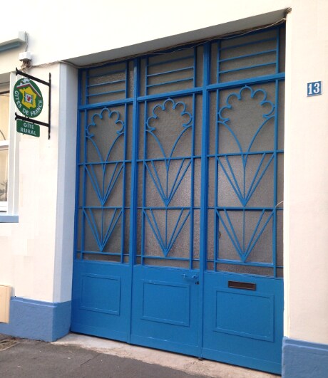 A vibrant blue double door features intricate geometric designs, enhancing the entrance of the Gîte Bleu. The doors are framed by a light-colored wall and a sign hangs above, indicating the property name. The overall appearance is bright and inviting.