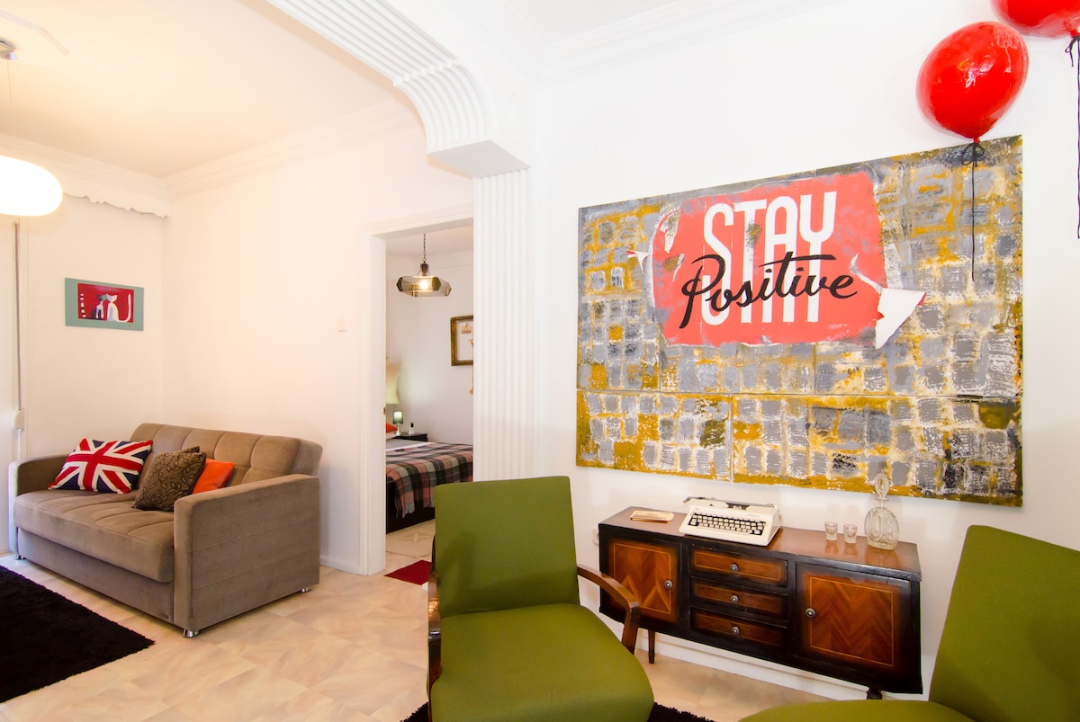 A well-decorated living area is presented, featuring a gray sofa paired with vibrant cushions. Two green chairs are positioned in front of a retro-style wooden console adorned with decorative items. A large, colorful artwork with the phrase 'STAY Positive' serves as a focal point, enhancing the inviting atmosphere.