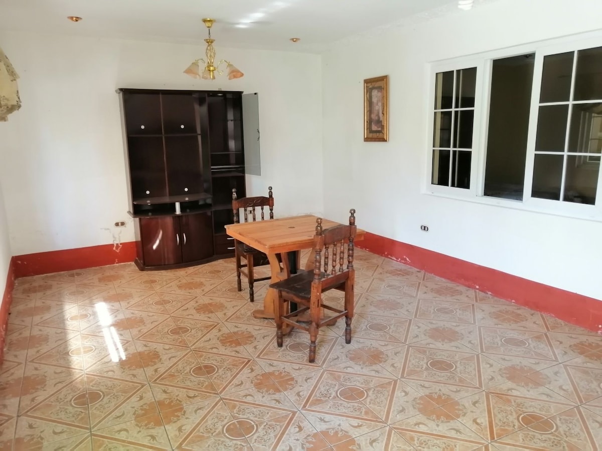 A simple dining area is presented with a wooden table surrounded by two chairs. A dark wooden cabinet occupies one wall, while a framed artwork hangs nearby. Natural light enters through multiple windows, illuminating the tiled floor with decorative patterns.
