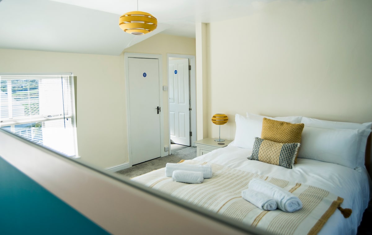 A bright sleeping area features a spacious bed with crisp white linens and a mustard yellow accent pillow. Soft towels are neatly arranged at the foot of the bed. Natural light fills the room through large windows, complemented by a decorative light fixture on the ceiling.