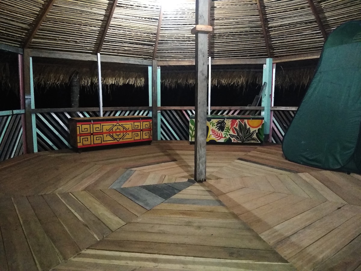 An open wooden structure features a hexagonal design with a bamboo ceiling above. Colorful wall art and a large decorative trunk add character. The wooden flooring showcases a unique pattern. Natural light from overhead illuminates the space, revealing the inviting atmosphere.
