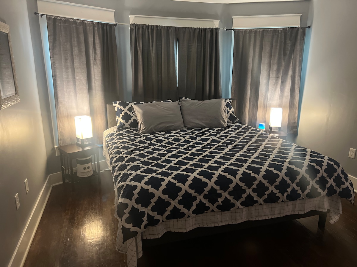 A spacious bedroom features a large bed covered with a geometric patterned quilt and two soft pillows. Natural light streams in through two windows, flanked by dark curtains. Two bedside lamps provide a gentle glow, adding to the room's warmth and comfort.