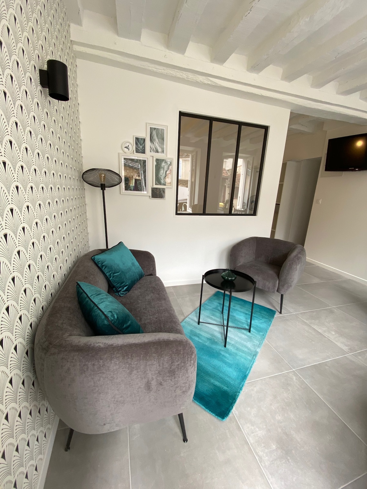 A cozy seating area is featured, furnished with two plush grey chairs adorned with teal cushions. A small black table rests on a matching teal rug, complementing the overall decor. A decorative wall with a patterned design enhances the space, while a window allows natural light to filter in.