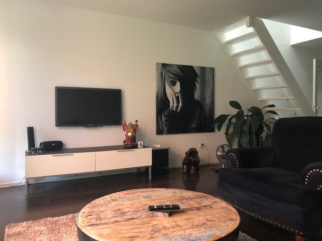 A spacious living room is characterized by a large, circular coffee table and a comfortable dark armchair. A television is mounted on the wall above a white cabinet. A striking black and white artwork is displayed, alongside lush indoor plants and a staircase leading to the upper level.