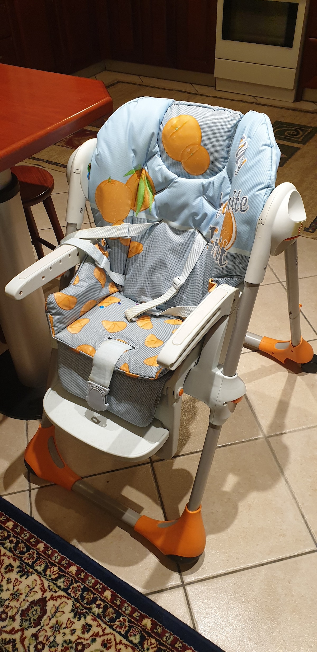 A high chair designed for young children features a padded seat with an orange fruit pattern. The chair is adjustable and includes a sturdy white frame, armrests, and a safety harness. Its base is equipped with non-slip feet for stability on tiled flooring.