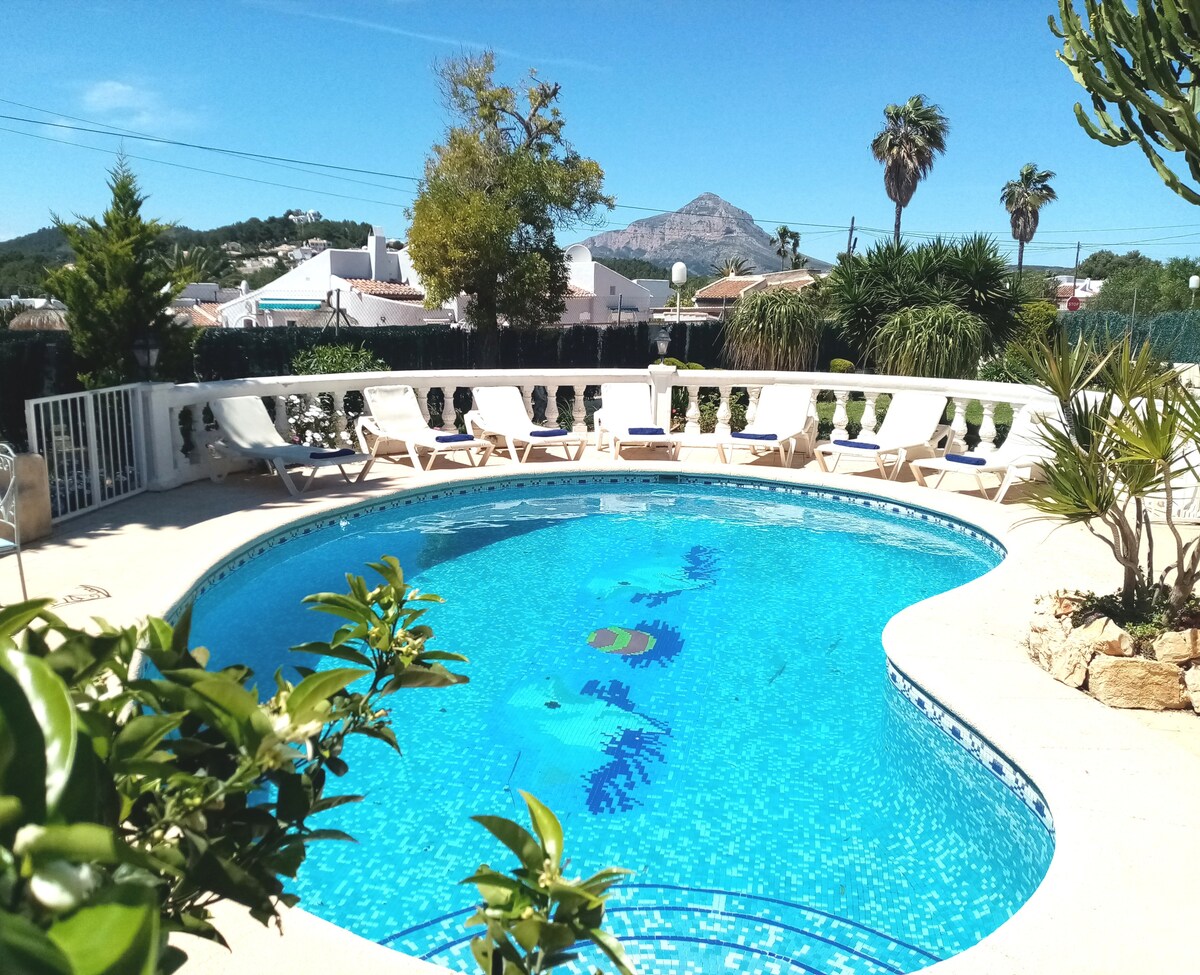A private swimming pool is surrounded by lounge chairs under clear blue skies. Lush greenery and palm trees enhance the scenic view, while a mountain rises in the background. The area is secured with a fence, ensuring a safe environment for guests.