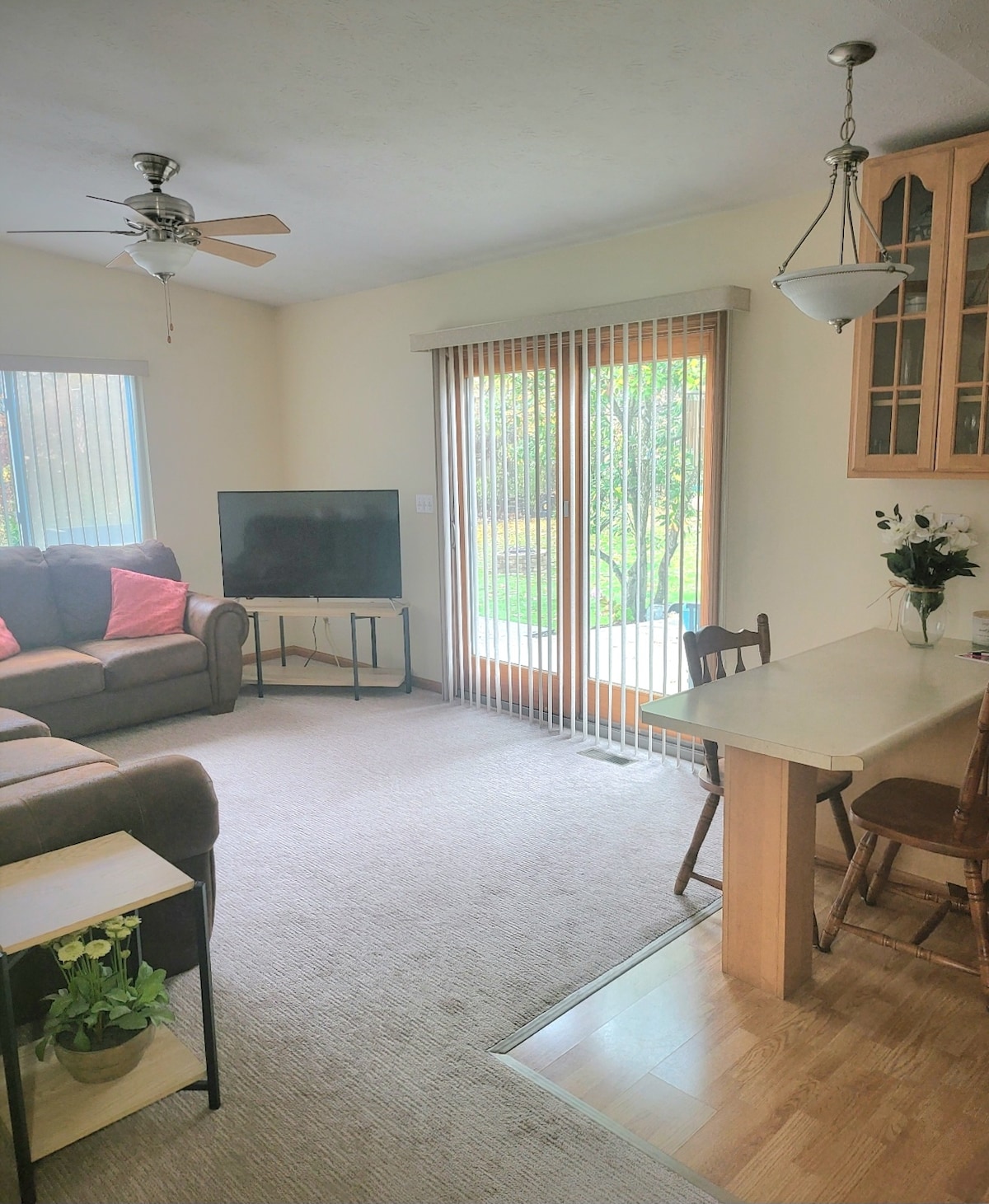 A cozy living area features a comfortable sofa set with orange accents and a small dining area. A TV is positioned on a stand, and sliding doors provide views of the outdoor space. Natural light filters through the blinds on the windows.