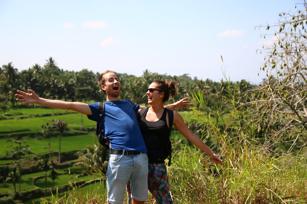 Two individuals are posed outdoors against a backdrop of lush green hills and palm trees. The scene captures an open landscape under a clear blue sky, with tall grass swaying gently in the foreground.
