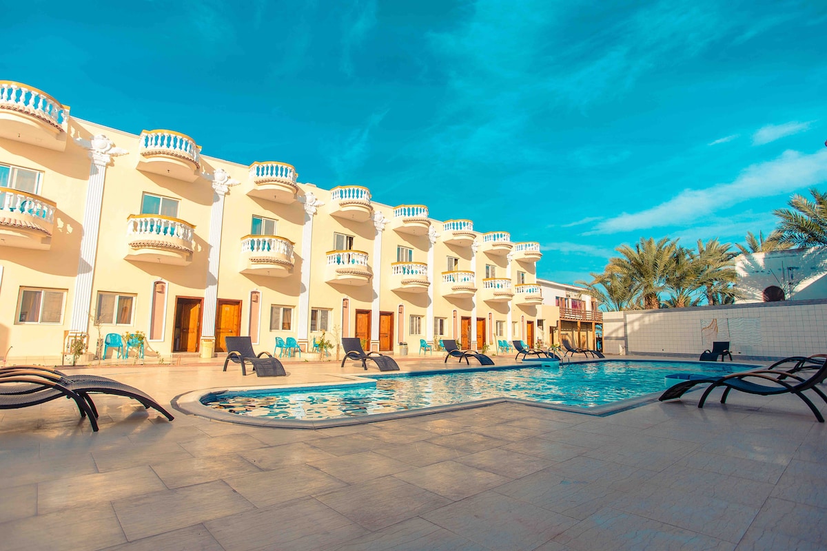 A rectangular swimming pool is surrounded by lounge chairs, providing a relaxing space for guests. The hotel structure features multiple balconies with white railings, offering a view of the pool area. Palm trees frame the scene, contributing to the inviting atmosphere.