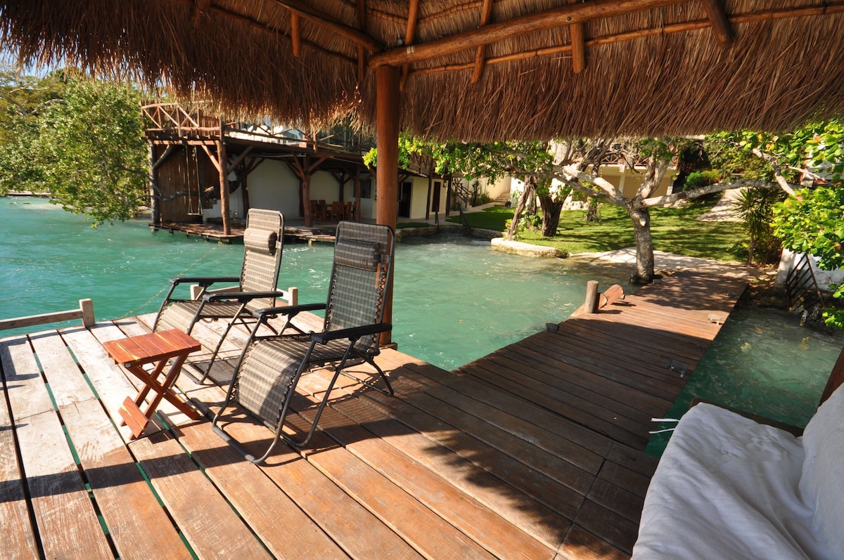 A wooden dock features two reclining chairs and a small side table, surrounded by calm turquoise waters. Lush greenery and a thatched-roof structure are visible in the background, creating a serene outdoor space for relaxation.
