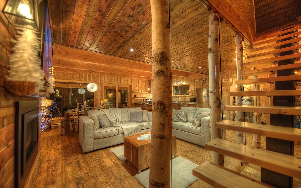 A spacious living area features wooden walls and ceilings, creating a warm atmosphere. A large, light gray sofa is centered around a wooden coffee table. Natural light enters through expansive windows, and a staircase with wooden steps leads to the upper floor.