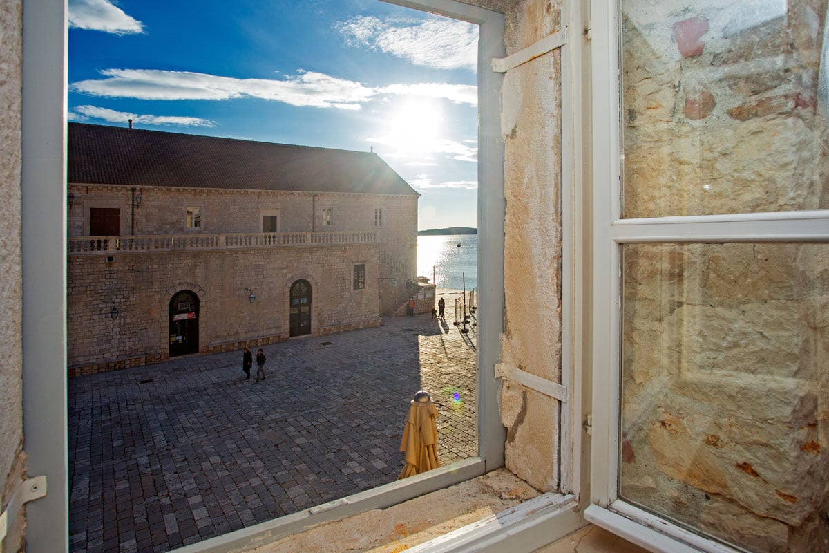A view from the apartment window captures the sun setting over the waterfront, illuminating the central square and showcasing stone buildings. A couple can be seen walking along the square, while a large umbrella is positioned nearby, enhancing the outdoor atmosphere.