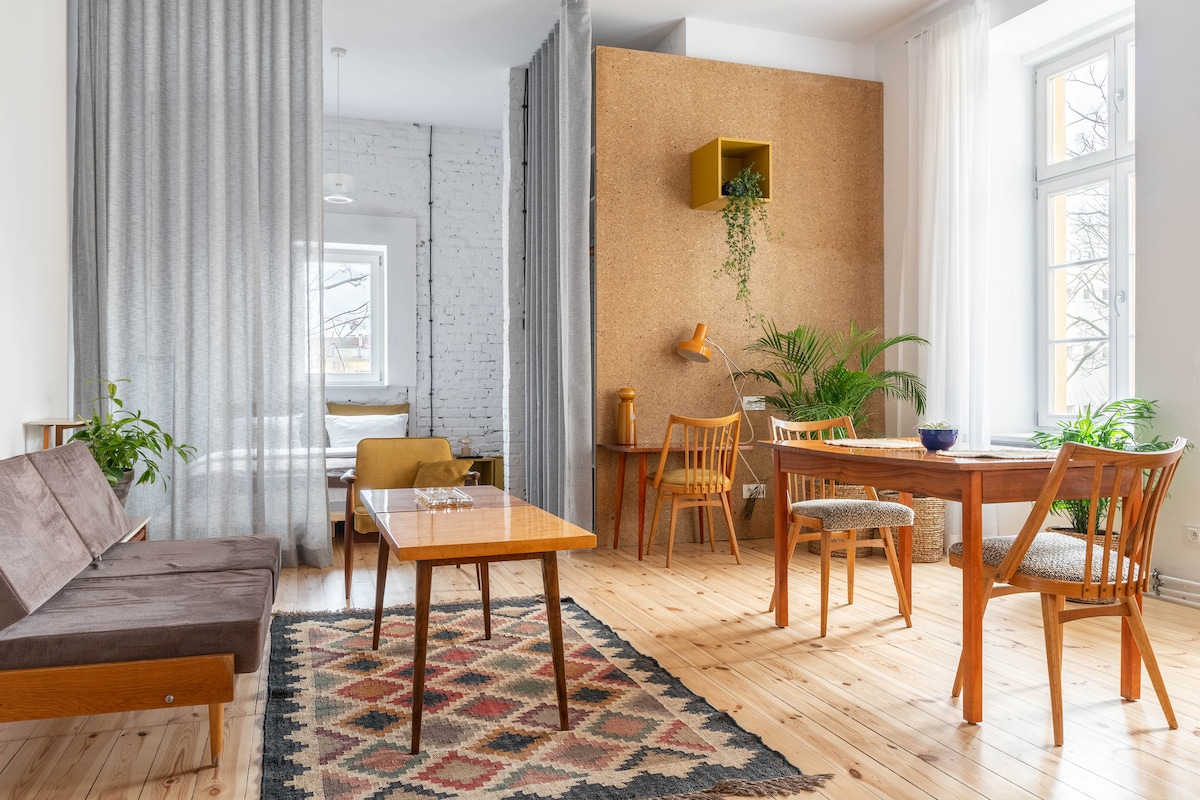 A bright and airy living space is divided by sheer curtains, featuring a mix of vintage wood furniture. A wooden dining table with chairs is positioned next to a small table, while plants provide a touch of greenery. Natural light enters through large windows.