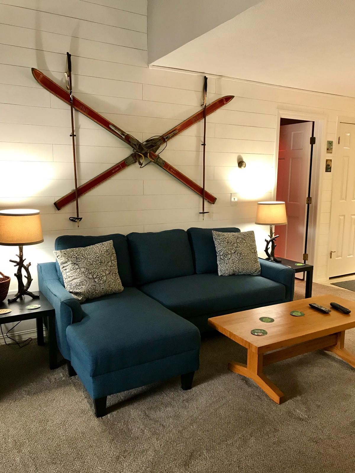 A cozy living area is presented, featuring a blue sectional sofa complemented by decorative pillows. A wooden coffee table sits in front, with remote controls arranged on top. Vintage skis are mounted on the wall, adding a rustic charm, while lamps provide soft lighting.