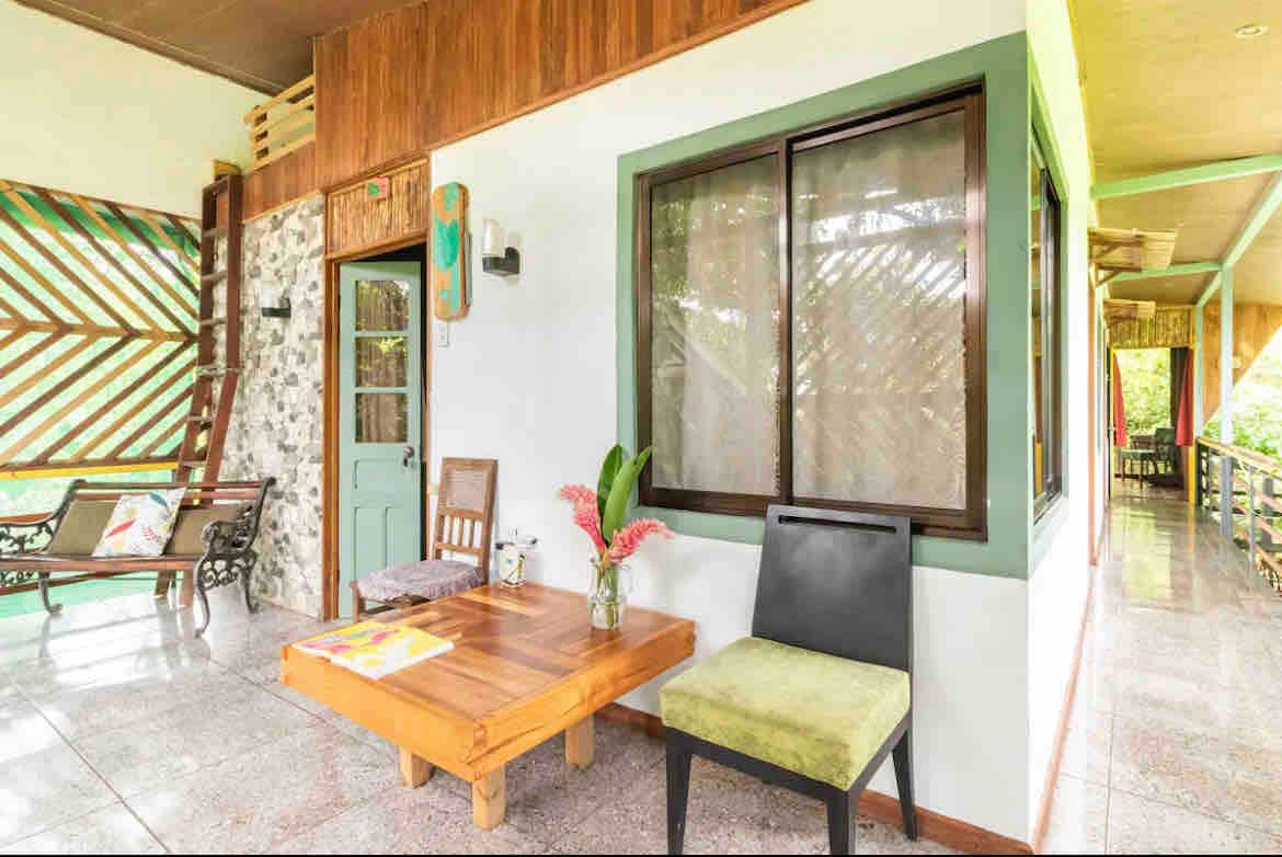 A welcoming open-air terrace is featured, showcasing a wooden table surrounded by a mix of seating options. Natural light filters through the surrounding greenery, and decorative elements add warmth to the space.