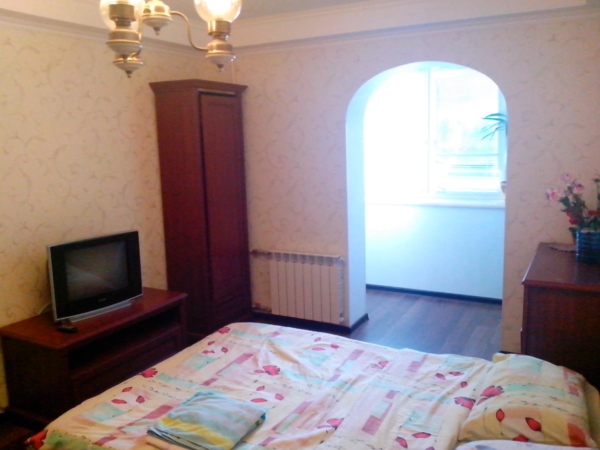 A well-appointed bedroom features a spacious bed with a colorful quilt. A wooden wardrobe and TV unit are positioned nearby. An archway leads to a bright area with a window, where natural light illuminates the space. Decorative plants and a floral arrangement add a touch of character.