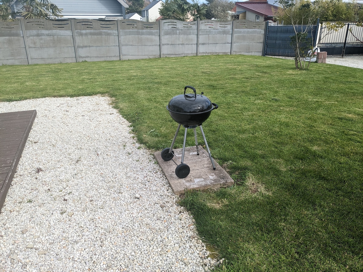 A well-maintained garden area is featured, showcasing a black barbecue grill positioned on a stone base. Lush green grass surrounds the grill, with a gravel pathway leading to a wooden deck visible in the foreground.