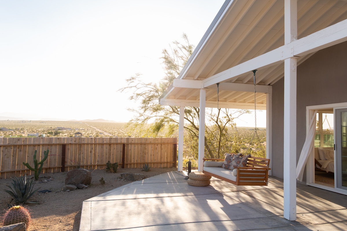 A welcoming porch features a comfortable swing, inviting relaxation while enjoying sweeping views of the desert landscape. Sunlight filters through the surrounding trees, casting gentle shadows on the concrete patio. Cacti and desert plants are visible in the yard, adding natural beauty to the setting.