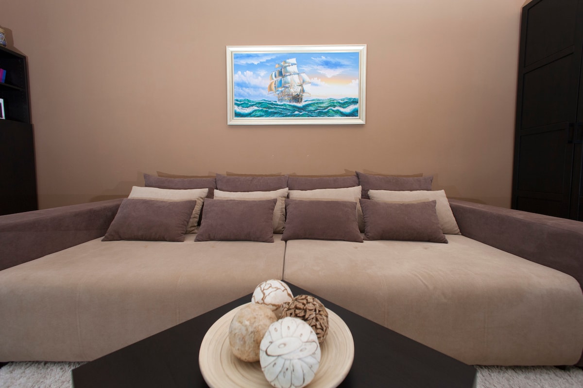 A spacious living area features a large, neutral-toned pull-out bed adorned with multiple soft cushions. A calming artwork of a ship sailing on water hangs above, while a round table in the foreground displays decorative natural elements.