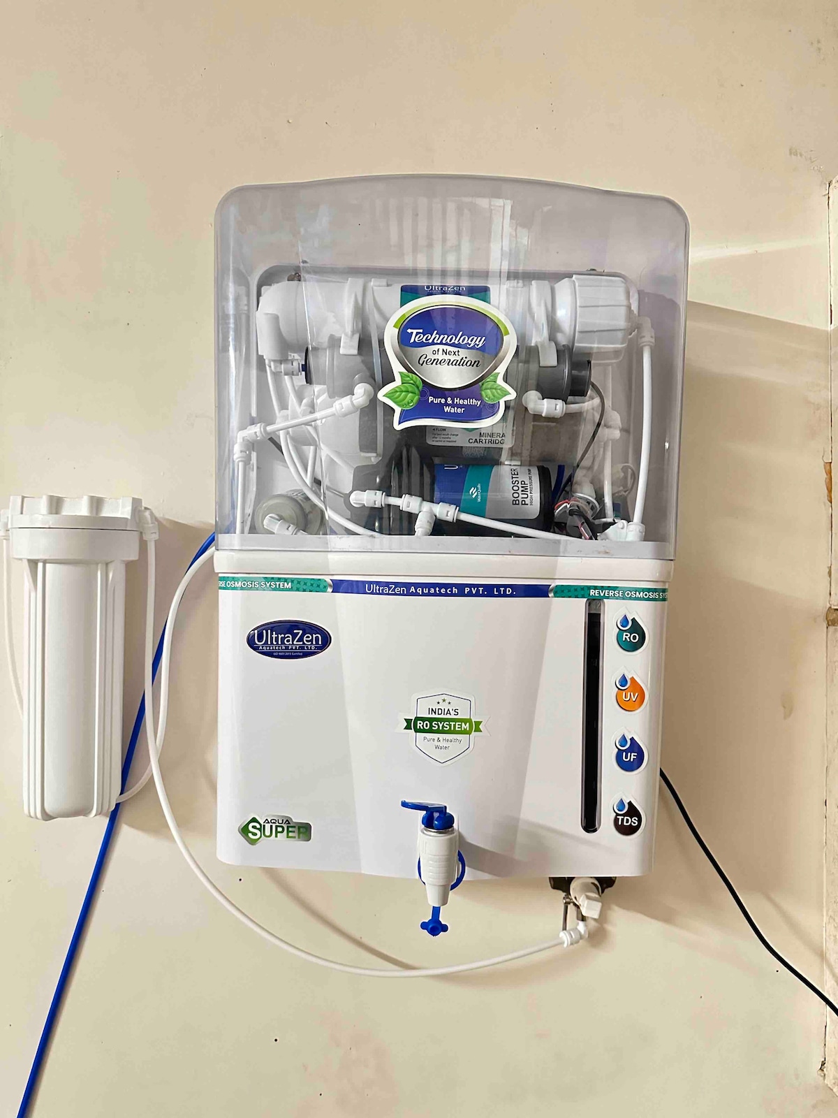 A water purification system is mounted on the wall, featuring a clear housing that displays several internal components. A blue water tap is positioned at the bottom, alongside filtration indicators on the front panel. The unit is surrounded by a simple wall finish.