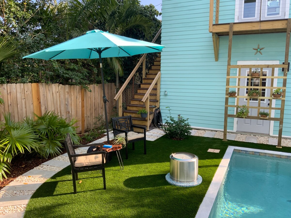 A private backyard area features a turquoise umbrella shading a cozy seating arrangement with two chairs. Lush green artificial grass surrounds a small decorative fire pit, with a saltwater pool partially visible in the foreground. A staircase leads to the upper level of the structure.