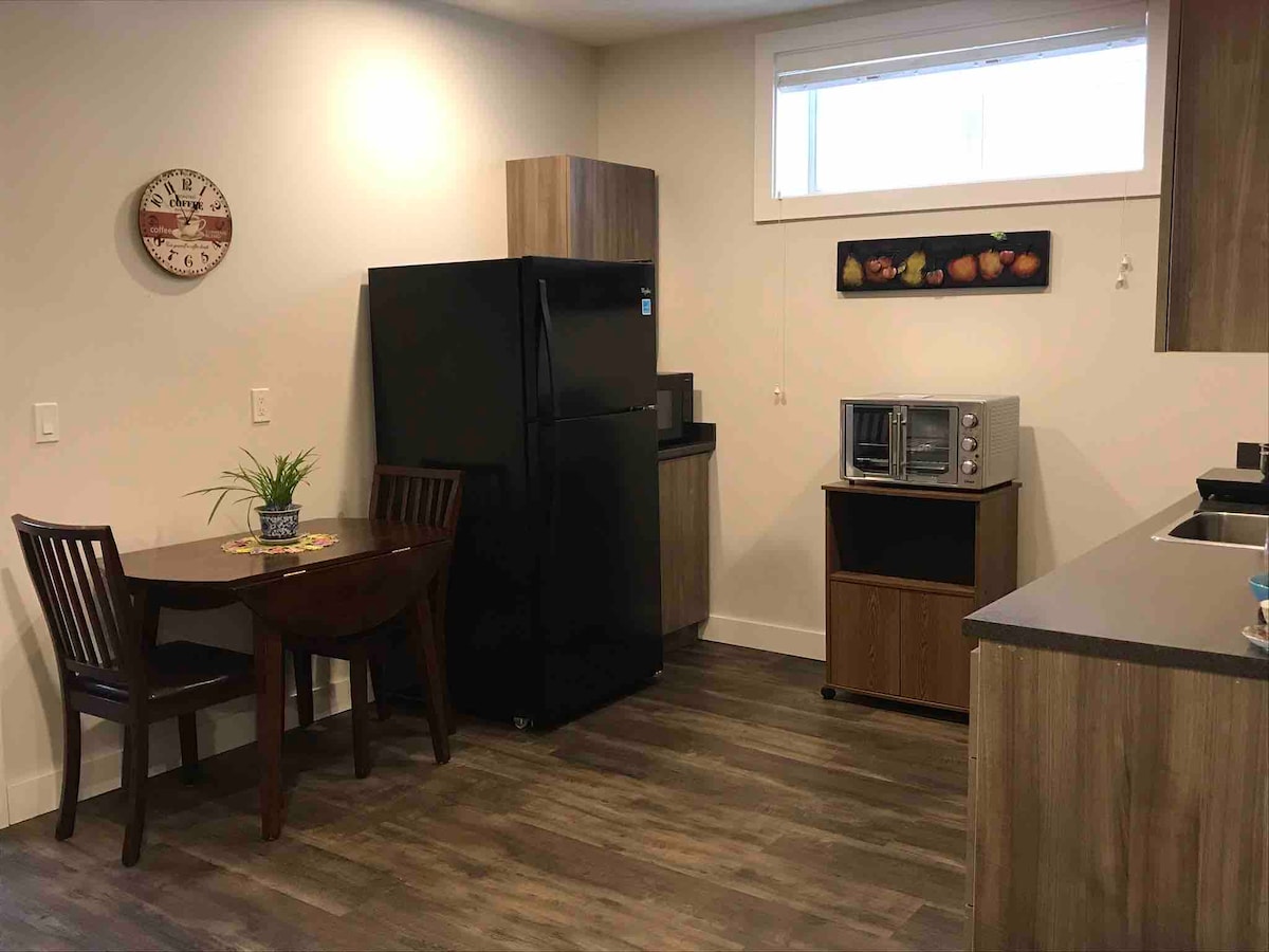 The kitchen area includes a black full-sized fridge and a microwave, along with a convection oven. A small wooden dining table with two chairs is positioned nearby. Natural light enters through a large window, illuminating the wood-patterned flooring.