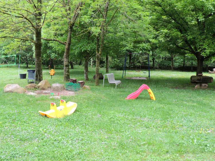 A spacious green area is surrounded by trees, featuring a small slide and a seesaw. A swing set is present in the background, along with a couple of chairs arranged on the grass. The open space is ideal for children to play.