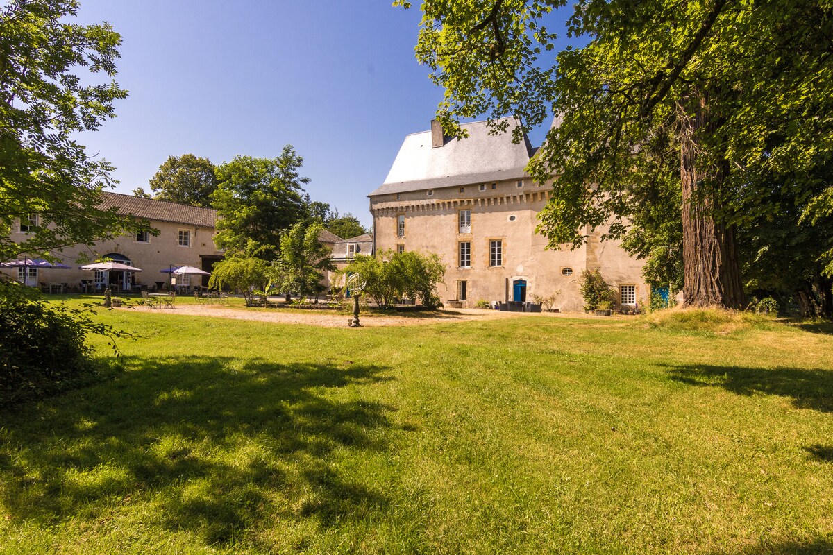 A landscaped area surrounds the château, featuring sprawling green lawns and mature trees. The building's façade is characterized by a sloped roof and large windows. Outdoor seating areas are visible, inviting relaxation in the open air.