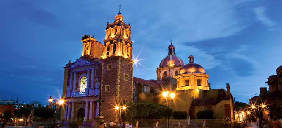 Illuminated colonial buildings are showcased against a twilight sky, highlighting intricate architectural details. The prominent church features a vibrant stained-glass window, while the neighboring dome reflects a soft glow, enhancing the charm of the historic square.