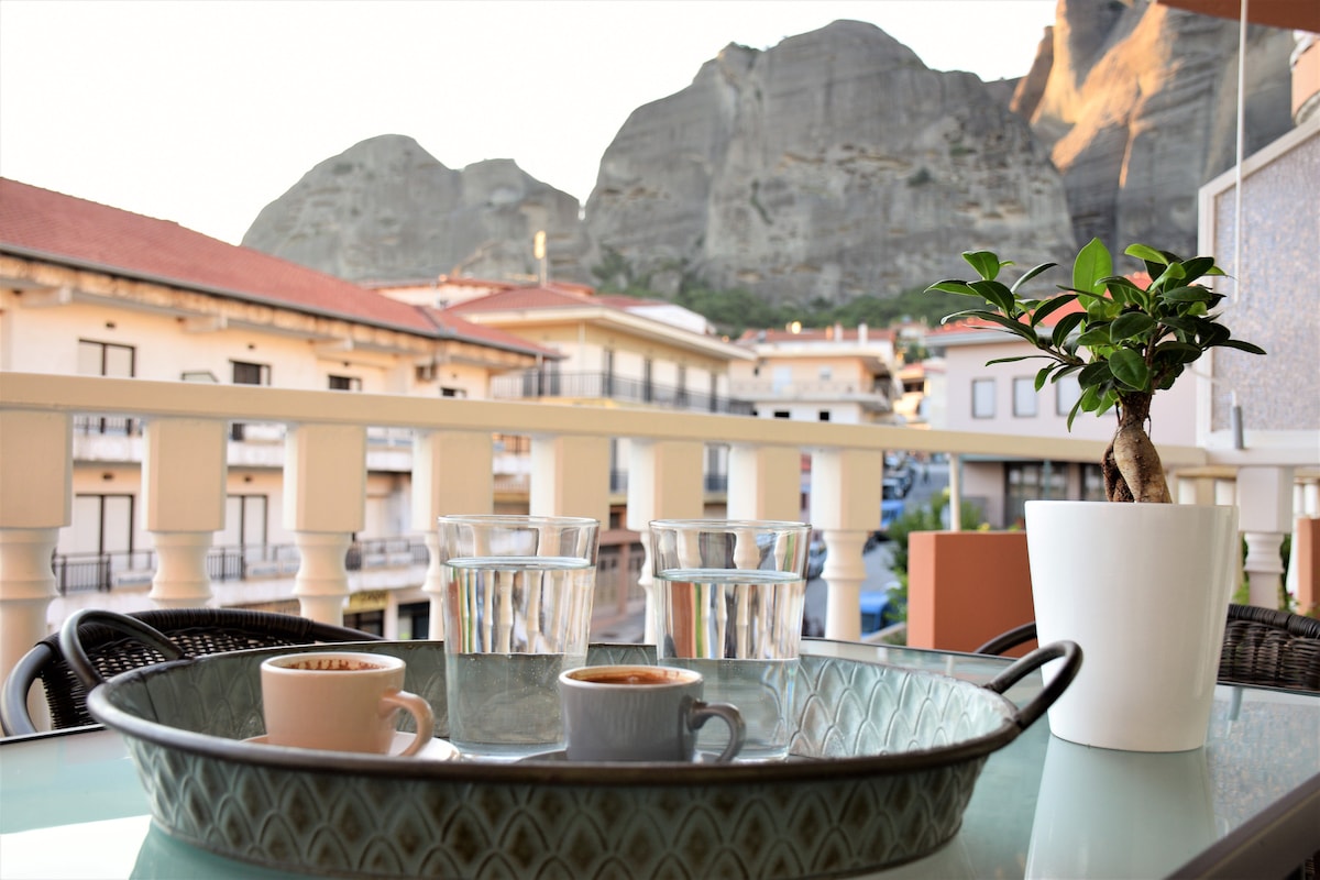A balcony view showcases a tray with two espresso cups and glasses of water, set against the backdrop of towering rocks. A small potted plant adds a natural touch to the table, while the surrounding buildings are visible in soft focus.