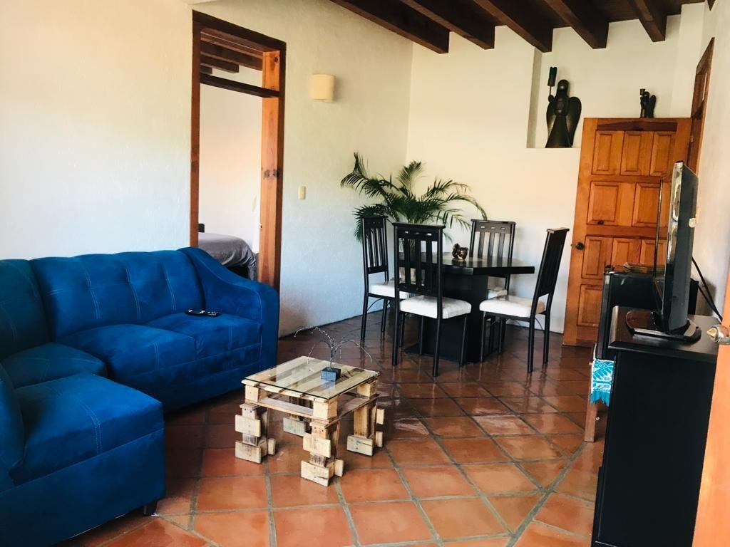 A living area features a blue sofa, a wooden coffee table, and a dining table with six chairs. Natural light illuminates the space through ceiling beams, and a plant adds a touch of greenery. A doorway leads to another room in the background.