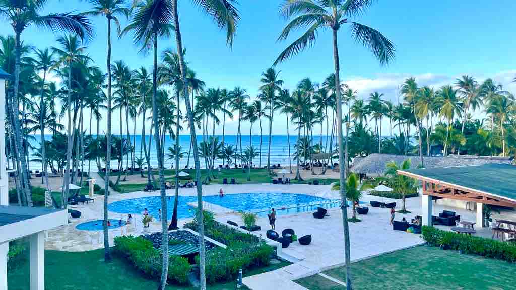 A serene view showcases a sparkling pool bordered by lounge seating and palm trees, with the beach and ocean visible in the background. The landscape includes well-kept grassy areas and a variety of seating arrangements, promoting relaxation and outdoor enjoyment.