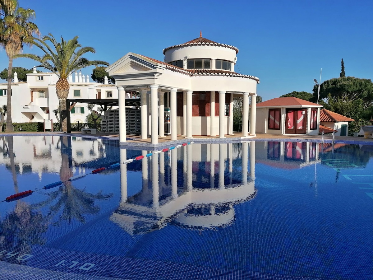 A modern architectural structure stands adjacent to a large swimming pool, reflecting its design in the clear water. Palm trees are visible in the background, complementing the vibrant blue sky. The pool area features lanes marked by ropes, enhancing its recreational aspect.