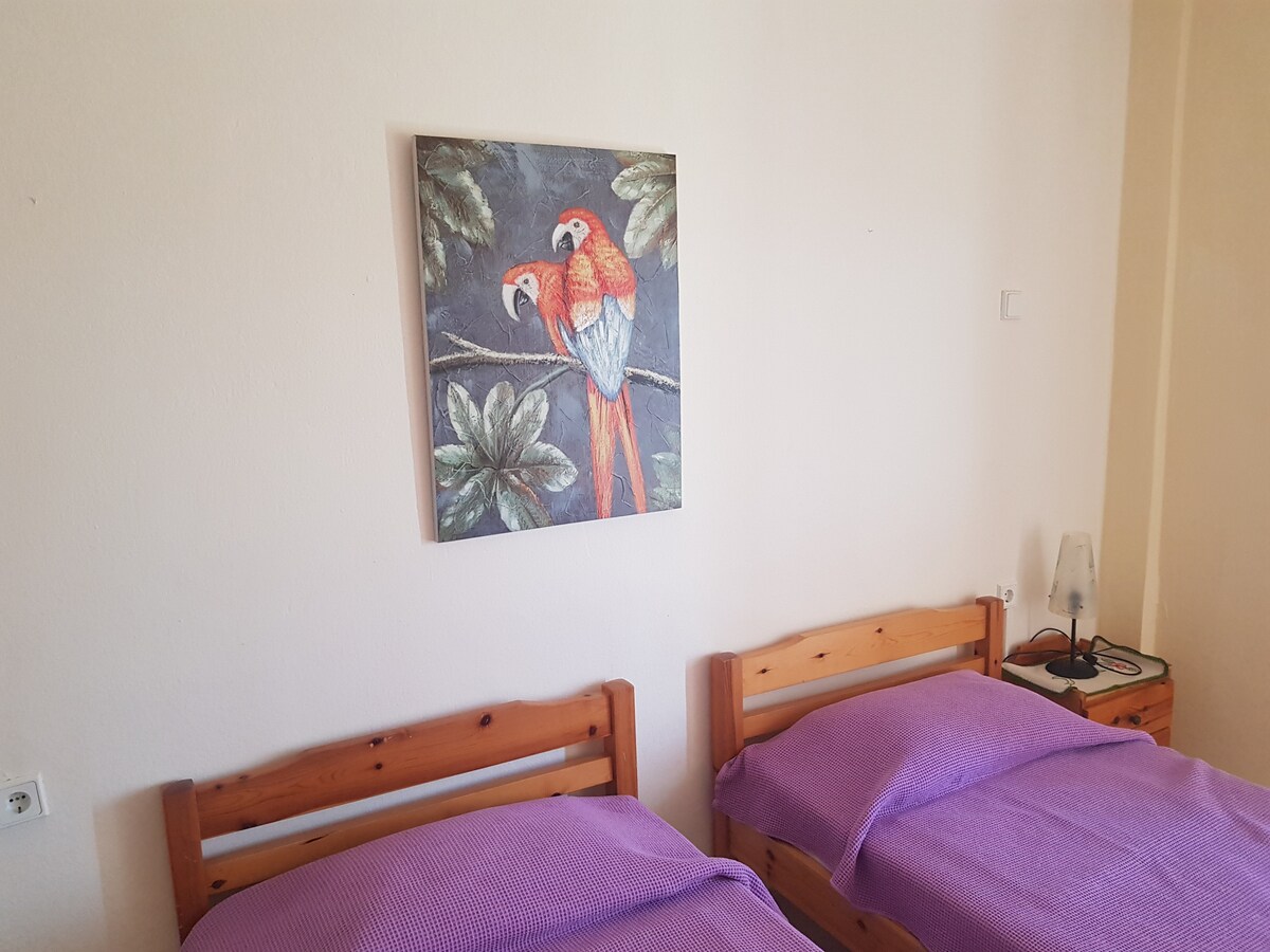 A cozy bedroom features two wooden beds covered with purple blankets. A bedside table holds a lamp, and a colorful painting of parrots hangs above the beds, surrounded by leafy greenery that adds a touch of nature.