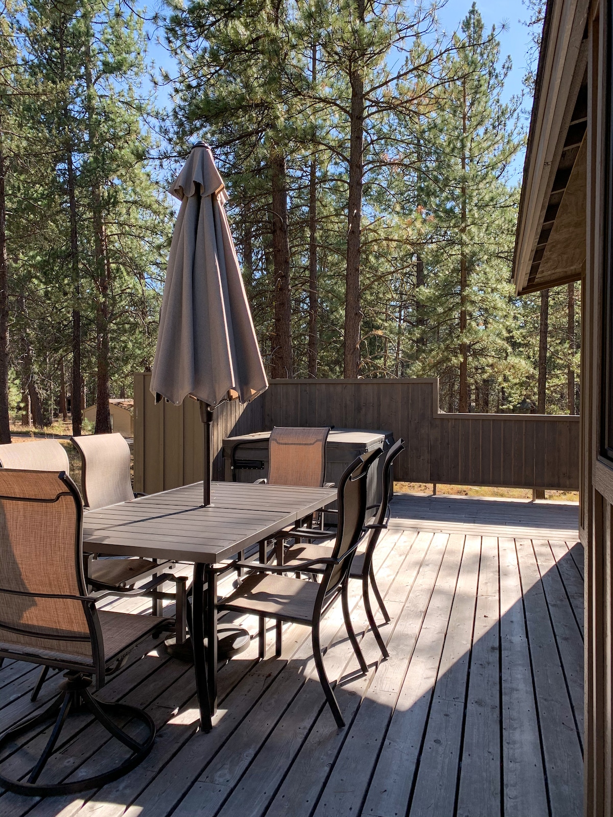 A spacious outdoor deck is shown with a dining table and several chairs arranged for seating. A large umbrella provides shade, while a BBQ grill is positioned nearby. The deck is surrounded by tall trees, creating a peaceful, natural setting.