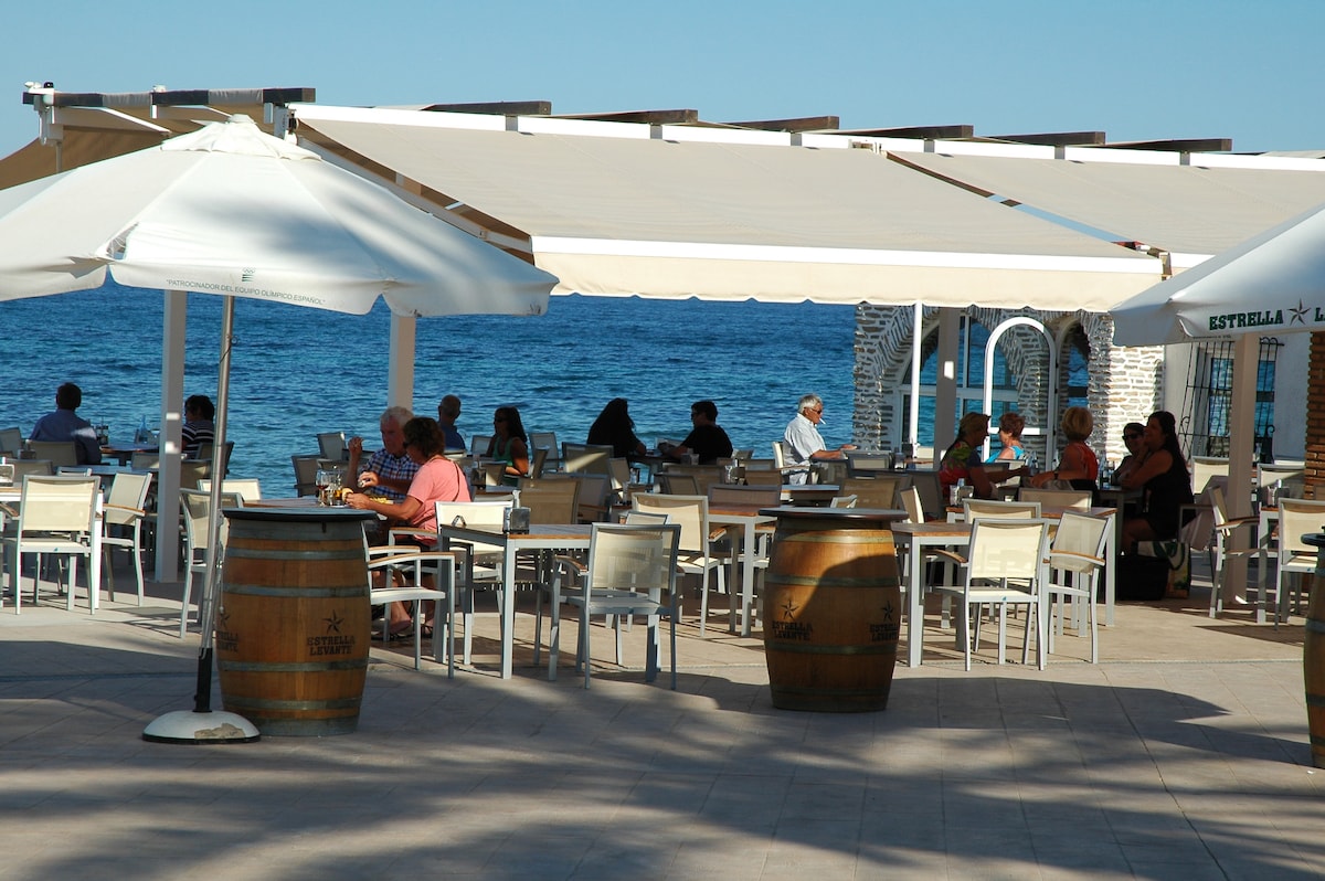 A spacious outdoor dining area is outlined by white umbrellas, providing shade to patrons seated at tables. Wooden barrels are placed decoratively around the area, with views of the sea visible in the background. The atmosphere appears relaxed, with several groups enjoying casual meals.