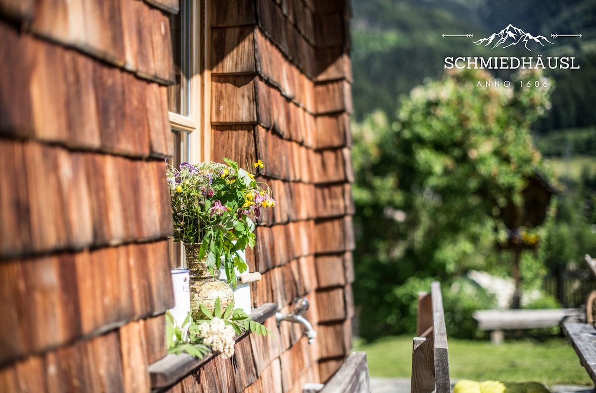 A wooden exterior showcases traditional shingle siding with a flower box filled with colorful blossoms beneath a window. A rustic table and benches are present in the foreground, accompanied by a natural backdrop of greenery and mountains, inviting a connection to the surrounding landscape.