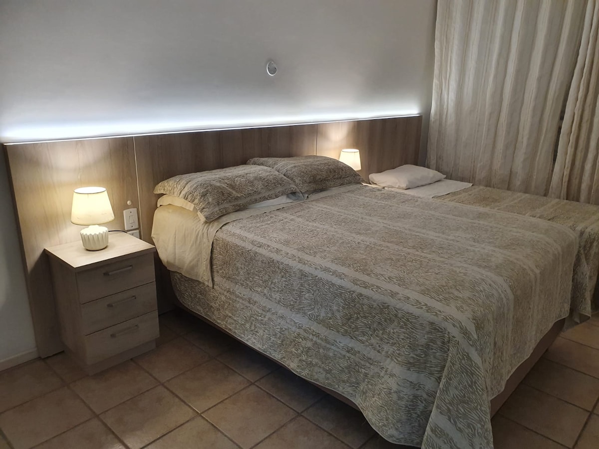 A well-appointed sleeping area features a spacious bed with a soft, patterned coverlet and a single bed beside it. Soft lighting emanates from two bedside lamps, while light curtains frame the window, allowing ambient light to enhance the calm environment.