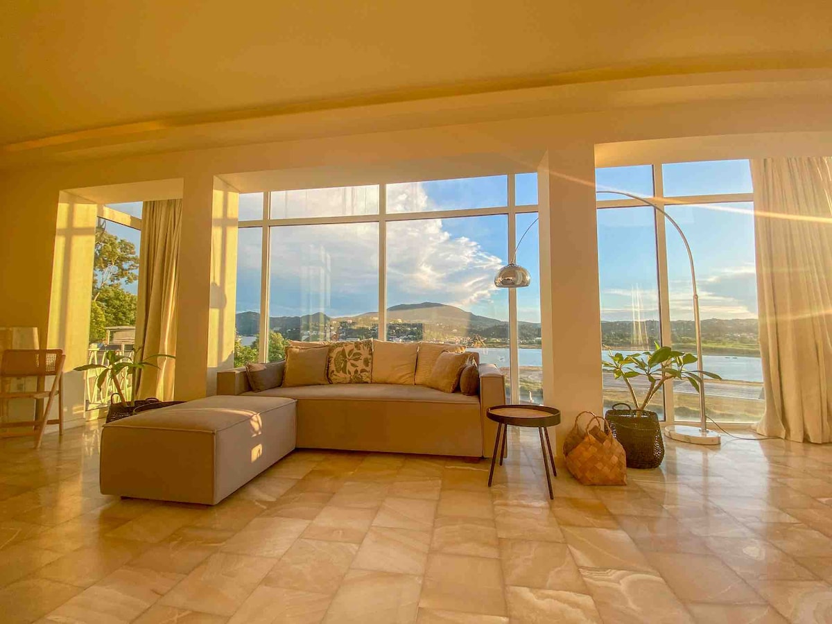 A spacious living area features large windows offering expansive views of the sea and mountains under a vibrant sky. A sleek gray sofa is complemented by a floor lamp and greenery, adding to the natural light illuminating the warm-toned flooring.